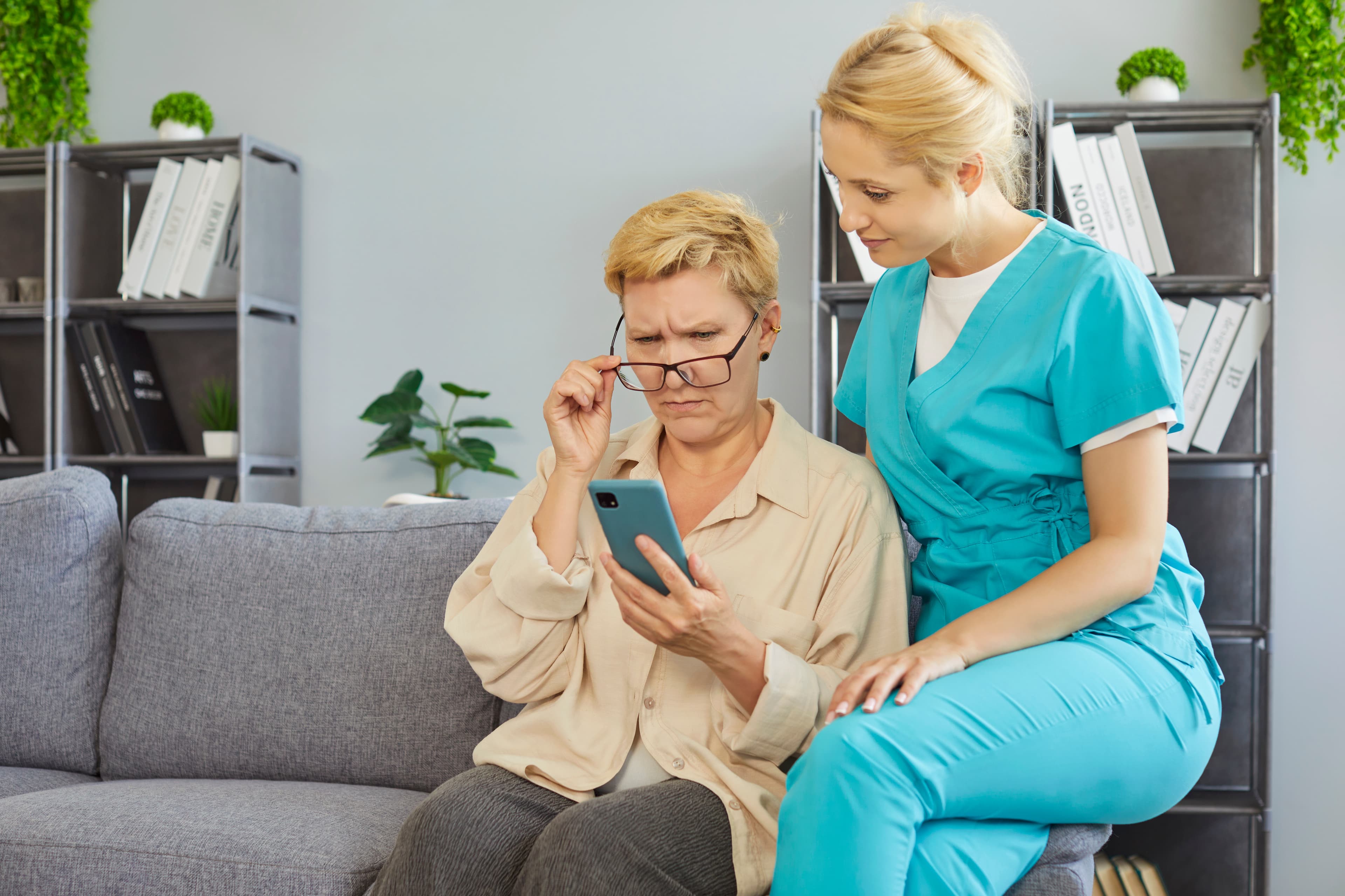 An older woman and a nurse in teal scrubs sitting on a couch, looking at a phone together